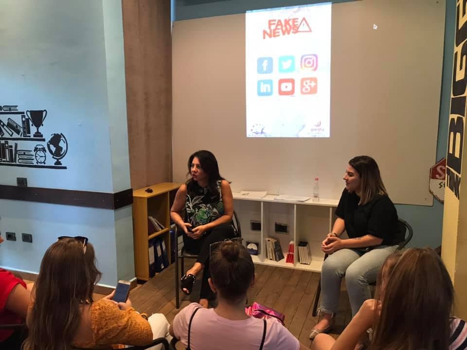 Folëse duke diskutuar me pjesëmarrësit; pas saj një raft librash dhe ekran me temën e takimit.