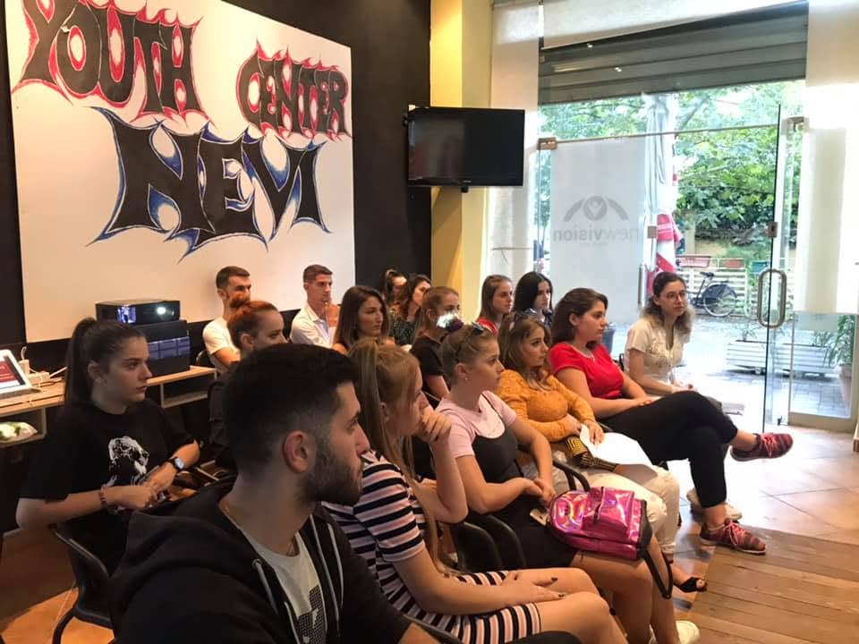 Të rinj të ulur në një sallë, duke ndjekur një takim; në mur një pikturë me mbishkrimin “Youth Center Nevi”.
